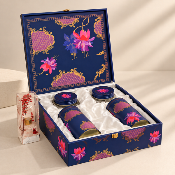 An open navy blue gift box with a floral gold and pink pattern, containing three matching cylindrical jars with lids, plus a small decorative glass vial placed beside it on a light beige surface.